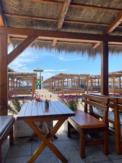 The image depicts an outdoor seating area, likely a restaurant or cafe, situated in Ulcinj, Montenegro. The scene is framed by a wooden structure with a thatched roof, suggesting a beachside or coastal setting. Rows of beach umbrellas and lounge chairs extend into the distance, indicating a popular tourist destination. In the foreground, wooden tables and benches with tan cushions are arranged for patrons. Potted plants with pink flowers add a touch of color. The sky is clear and blue.