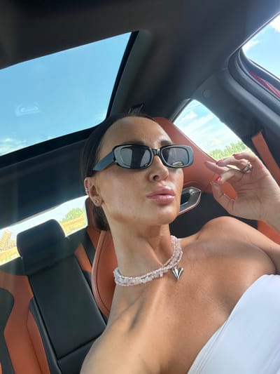 This media file shows a woman inside a car, with the sunroof open, allowing natural light to stream in. The woman is wearing sunglasses and a white top. She has a delicate necklace with a heart pendant and a bracelet made of pink stones. Her fingernails are painted red. The car's interior appears to be luxurious, with leather seats in shades of black and brown. The sky is visible through the sunroof, suggesting it is daytime. There is no visible text in the image. The setting appears to be Zapadny, Russia, based on the provided location context. The overall mood conveyed is one of relaxation and enjoyment of a sunny day.