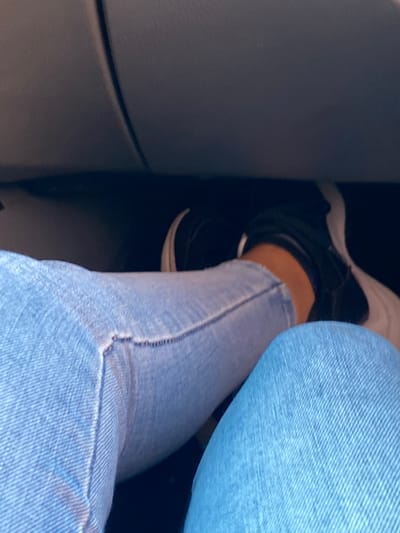 The image captures a close-up, low-angle view of a person's legs and feet inside what appears to be a vehicle, possibly a car, in La Cruz del Pastel, Venezuela. The subject is wearing light blue denim jeans with visible stitching and texture, and black and white athletic sneakers. One foot is more clearly visible, showing a black upper with white soles and a glimpse of an orange-brown ankle. The person's legs are bent, indicating they are seated.

The background consists of dark, likely molded plastic or fabric interior surfaces of the vehicle, which are out of focus and provide a deep, shadowed contrast to the lighter clothing. The lighting is somewhat dim, suggesting an indoor setting or a shaded environment within the vehicle. No specific activity beyond being seated is evident. There is no visible text in the image.