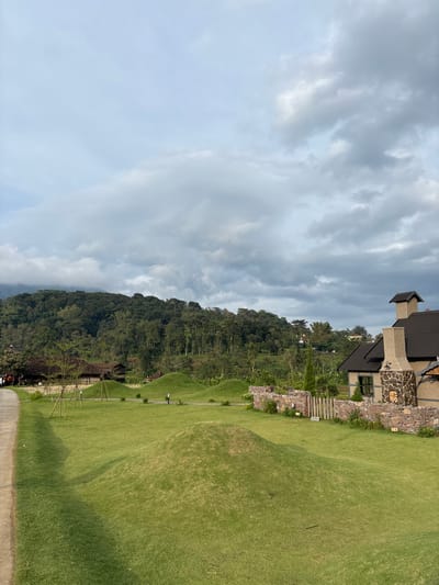 This image captures a wide outdoor landscape in Tamiajeng, Indonesia, under a sky densely covered with light and dark grey clouds. The foreground features a manicured green lawn with multiple smooth, rounded, artificial grass mounds of varying sizes scattered across it. A narrow, light-colored paved pathway runs along the far-left edge, ascending slightly towards the background. In the midground to the right, a building with a dark, sloped roof and light-colored walls is visible, featuring a prominent, tall exterior chimney constructed from irregular stacked stones; an opening within the chimney contains cut firewood. Adjacent to this building is a stone wall, also made of irregular masonry, incorporating a small wooden gate. Beyond the lawn, in the left-midground, several traditional-style wooden structures with dark roofs are partially obscured by sparse trees. The background is dominated by a dense, dark green forest covering a large hillside, extending across the entire horizon and meeting the cloudy sky. No discernible individuals or explicit actions are present within the scene.
