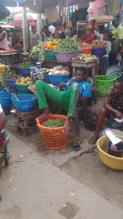 In Warri, Nigeria, a bustling marketplace is captured. A young man in a blue shirt and green pants reclines in a plastic chair, his bare feet resting in a filled orange plastic basket. Next to him, a woman sits on the ground using a knife to peel vegetables and dropping them into a yellow bucket. Surrounding them are multiple baskets filled with vegetables and fruits. Other people are visible in the background, along with various vendors and market stalls.