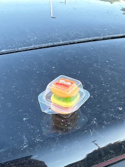 The image shows a small, clear plastic container with a label that reads "Trolli Original" and some gummy candies inside. The container is resting on a dark, dusty surface, likely the hood of a car. The candies appear to be fruit-flavored, with visible layers of orange and green. The background is out of focus but suggests an outdoor setting, possibly with a reflection of the sky and some distant structures. The lighting indicates it could be daytime, but the exact time is not discernible. The presence of dust on the surface suggests an environment that is not pristine. There are no people visible in the image. The overall scene is a close-up of an object on a textured surface, with no apparent activity taking place. The location context provided is Bagard, France, but no specific visual cues in the image confirm this.