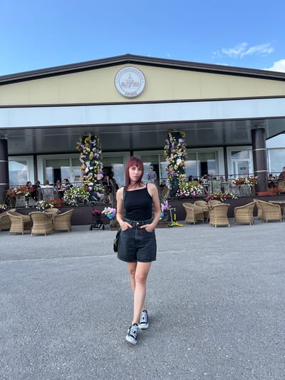 A young woman with red hair stands in the foreground facing the camera, hands in her pockets. She is wearing a black tank top and denim shorts, with black and white high-top sneakers. Behind her, a building with a light yellow facade and a dark brown roof features a large sign with a circular emblem. The emblem contains an illustration of a building and text in Russian. The building has a covered outdoor seating area with wicker chairs and tables, decorated with numerous flower arrangements in baskets and planters. Several people are seated at the tables in the background. The ground in front of the building is asphalt, and the sky above is blue with scattered clouds. The location is identified as Akhlebaikha (Kovriginskii S/S), Russia.