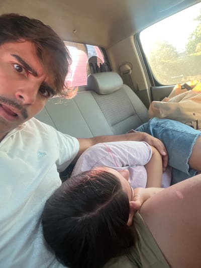A car interior shows a person in a white shirt taking a selfie. A woman in a pink shirt rests next to him, seemingly asleep. Light blue denim shorts are also visible. The car seat is light grey. The scene is bright due to sunlight filtering through the car windows. The driver appears to be looking directly at the camera. The picture is angled, presenting an unconventional perspective.