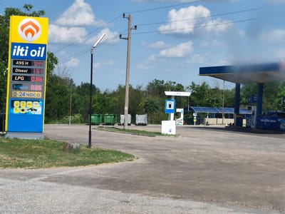This is a daytime, eye-level, medium-shot photograph of a gas station in Totleben, Bulgaria. A prominent yellow sign on the left displays the "ilti oil" logo and current fuel prices: A95 H at 1.44, Diesel at 1.51, and LPG at 0.66. It also indicates AdBlue at 0.85 and that the station is open 24 hours, with icons for a store, Wi-Fi, restrooms, and payment options. In the background, a blue and yellow gas station canopy with pumps is visible. A blue car is parked at one of the pumps. To the left of the canopy, there are several green and silver trash bins. A utility pole with wires runs across the sky. The ground is paved with asphalt, and the area is surrounded by green trees under a partly cloudy blue sky.