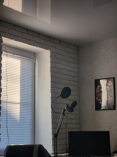 The image depicts an indoor scene, likely a home office or recording studio, in Murom, Russia. A window with white blinds is visible on the left, letting in sunlight that casts striped shadows. To the right of the window, a white brick wall is partially visible. A microphone on a stand is positioned in the center of the frame. Further to the right, on a textured grey wall, a framed picture hangs, featuring a collage of portraits. Below the picture, a black computer monitor is placed on a surface. A black office chair is partially visible in the lower left corner. The ceiling is a glossy white surface with reflections. The overall lighting suggests it is daytime.