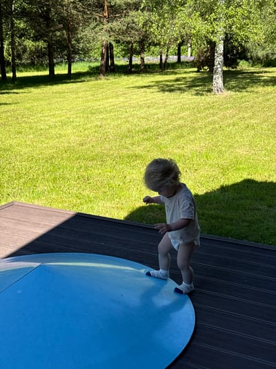 The image depicts an outdoor scene in Копнино, Russia, on a bright, sunny day. A young child, appearing to be a toddler with light blonde, curly hair, is positioned towards the lower center-right. The child is wearing a light-colored short-sleeved t-shirt, a white diaper, and white socks with blue and red stripes. The child's gaze is directed downwards, towards their feet and a large, round, blue object on the ground.
The child is standing on a dark grey or brown composite deck with horizontal grooves. The right foot of the child is partially placed on the blue object, which appears to be a flat or slightly domed cover, possibly for a trampoline or a small pool, flush with or resting on the deck surface. A seam or fold is visible across the blue surface.
Beyond the deck and the child, a vibrant green, neatly mown lawn extends across the mid-ground. In the background, a dense line of trees forms a natural boundary. These include tall pine trees with dark trunks and green needles, as well as a prominent birch tree with a distinctive white trunk and bright green leaves on the right side of the background. Hints of purple vegetation or flowers are discernible amongst the trees in the distance. The sunlight casts distinct shadows on the deck from an unseen object to the left, and shadows from the trees are visible on the lawn, indicating a clear, sunny sky.