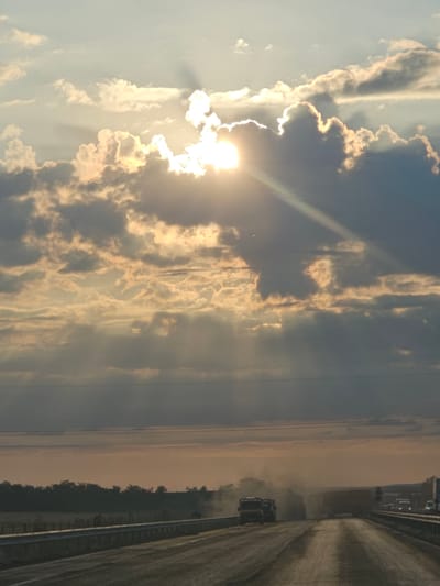 The image depicts a highway scene at dusk in an unknown location in Bulgaria. The sky is dramatic with the sun partially obscured by dark clouds, casting god rays across the scene. Several vehicles, including trucks and cars, are visible on the road. One truck in the foreground is kicking up a significant amount of dust, indicating it might be a construction or agricultural vehicle. The road appears to be under construction or in a state of disrepair, with loose gravel and dirt. A guardrail lines the side of the road. The landscape beyond the highway consists of a line of trees and a flat, open terrain. Power lines are visible in the sky.