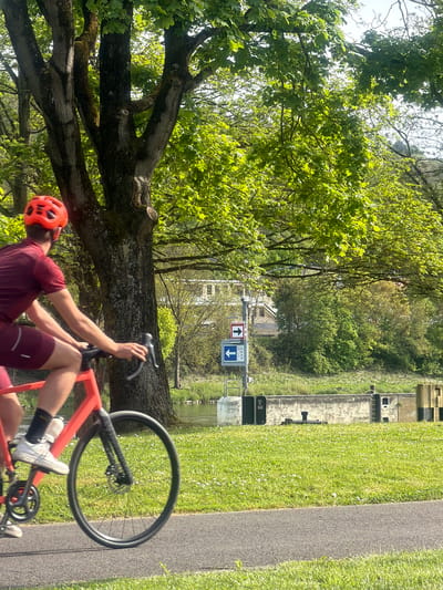 This image captures an outdoor scene in Grevenmacher, Luxembourg, on a bright, sunny day, likely during spring or summer. The primary subject is a male cyclist, seen resting or pausing on a vibrant red bicycle. He is wearing a red cycling helmet, a maroon short-sleeved jersey, and matching shorts, with white cycling shoes.
The setting features a canal or river on the right side of the frame, bordered by a well-maintained grassy bank and a paved path. In the background, concrete structures, indicative of canal locks, are visible along the waterway, suggesting its use for navigation. Lush green trees and bushes dominate the middle ground, with a large, mature tree featuring extensive green foliage in the left foreground.
In the mid-distance, two square signs are visible above the waterway. One is white with a prominent red upward-pointing arrow. The other is blue with a white downward-pointing arrow and the text "L.C.P. 15.00" displayed beneath it. The bright sunlight creates clear shadows and highlights the verdant landscape, contributing to a calm, recreational atmosphere.