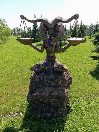 A statue of a figure resembling the astrological sign Libra is displayed outdoors in a park-like setting. The statue is dark brown and depicts a seated, female-like figure with large, curved horns or headdresses and a serene facial expression. She holds scales in each hand, with empty pans. The figure is seated on a rough, rock-like base. Inscribed on the base are the letters "BEGL" in a decorative font. The statue is positioned on a lawn of green grass, with scattered white wildflowers. In the background, there are numerous evergreen trees, some deciduous trees with green foliage, and a clear blue sky. The overall environment suggests a sunny day in a recreational area. The city of Il'ich, Russia, is mentioned as the location, implying this park is within that city.