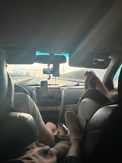 The image captures a rotated view from inside a vehicle, looking towards the front dashboard and windshield, under bright daytime sunlight. The setting appears to be a car journey or a stop along the way, possibly in Aleksandrovskoe, Russia.
Two people are visible: one person's legs and feet, clad in light-colored socks, are propped up on the right side of the car interior, suggesting a passenger relaxing. A crumpled snack bag, partially showing the Russian word "КАРТОШКА" (potato/crisps), lies nearby. Another person's hands and arms are seen closer to the steering wheel area on the left.
The car's interior features a dashboard with a central console displaying "8:50" and "88.2 FM2" on its screen. A rearview mirror is prominent, from which an "Areon X" branded air freshener hangs. Two smartphone mounts are attached, one holding a phone displaying a navigation map, and another phone is suspended near the rearview mirror. Sun visors are present, with one having its mirror cover open. Through the front windshield, a set of railway tracks runs parallel to the vehicle, suggesting the car is either parked next to or driving alongside a railway line. The bright light streaming in indicates a sunny day. The overall atmosphere appears casual and relaxed, indicative of travel.
