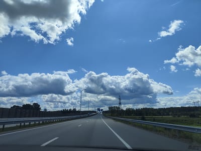 The image depicts a view from a vehicle traveling on a highway. The highway has multiple lanes with white lane markings. On the left side of the highway, there is a metal guardrail and a sound barrier wall. On the right side, there is also a guardrail and a grassy shoulder leading to a treeline. The sky is a vibrant blue with large, fluffy white clouds and some darker gray clouds. In the distance, there are trees lining the sides of the highway, and tall power line towers are visible. There are signs on poles along the highway. The scene appears to be located in a rural or semi-rural area. It is a bright, sunny day with some cloud cover. The location is identified as Katlakalns, Latvia.