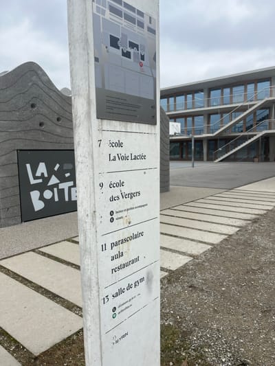 The image captures an outdoor scene at what appears to be an institutional or school complex in Meyrin, Switzerland, under an overcast sky during daytime. No people are visible.

In the foreground, a tall, light-colored concrete information pillar stands prominently. It features a map of the complex on a dark panel, alongside a directory with numbered listings. The directory includes: "7 école La Voie Lactée", "9 école des Vergers", "locaux en gestion accompagné", "scouts", "11 parascolaire", "aula", "restaurant", and "13 salle de gym". Towards the bottom of the pillar, the text "MEYRIN" is discernable, confirming the location context. Below the map, a dark rectangular sign, partially obscured by a textured concrete sculptural element, displays "LAX BOITE" in bold white letters.

The ground consists of a patterned paved walkway with rectangular concrete slabs, leading towards the background, and transitioning into a gravelly area on the right. In the midground and background, a modern, multi-story building with numerous large, horizontally oriented windows is visible. The building's glass facades reflect the grey, cloudy sky. The overall impression is one of a functional and well-organized public or educational facility.