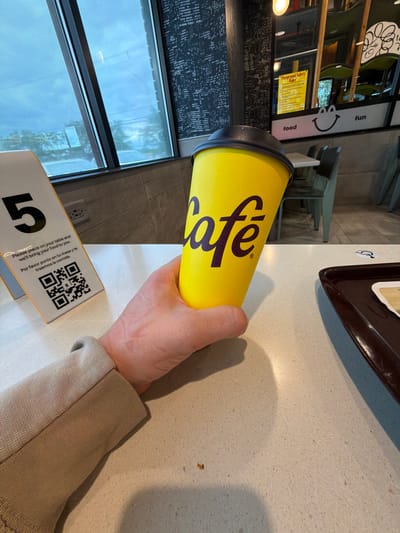 A person's hand holds a yellow cup with the word "Café" in purple lettering. The cup has a black lid and is positioned on a light-colored speckled countertop. To the left of the cup is a white table tent with the number "5" in black and English and Spanish text including a QR code. In the background, through a large window, a cloudy sky and trees are visible. The interior appears to be a restaurant, with tables and chairs, and a sign that says "Playground Safety Rules" in yellow. The overall impression is of a casual dining establishment, possibly a McDonald's given the yellow and purple branding and the mention of a playground. The location is Palatine, United States of America.