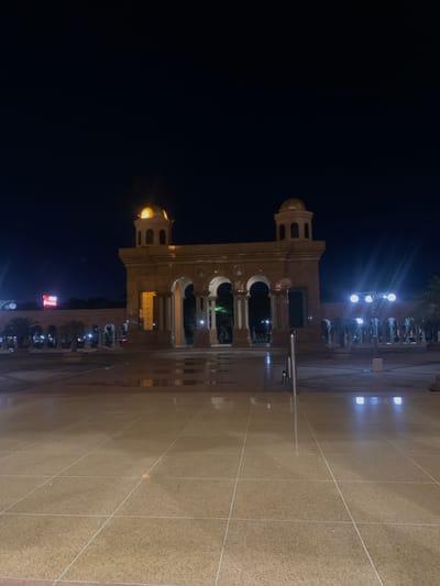 The image depicts a grand building illuminated at night, viewed from a slightly elevated perspective across a large, wet plaza. The sky is dark, indicating a late hour, and the scene is entirely lit by artificial sources, creating dramatic contrasts and reflections.

The central subject is an impressive architectural structure, possibly a mosque or a public building, featuring a warm, light-colored facade. It is characterized by two prominent towers, each topped with a golden dome that glows brightly under focused lights. The building incorporates numerous arched windows and doorways, some revealing glimpses of interior lighting or intricate patterns.

In front of the building, a vast, light-toned paved plaza stretches across the foreground. The surface appears wet, creating clear, shimmering reflections of the building's lights and the surrounding streetlights. White grid lines define the paving patterns on the ground. To the left of the main building, a cluster of bright streetlights with multiple fixtures casts strong beams, further illuminating the scene. Some dark foliage, likely trees, can be seen in the background, subtly lit by the streetlights.

No clear human figures or specific activities are discernible, giving the scene a quiet and contemplative atmosphere. The architectural style, with its domes and arches, is highly suggestive of Islamic design, aligning with the given location context of Samarinda, Indonesia. There is no visible text or identifiable signage in the image. The overall impression is one of serene majesty and striking nighttime illumination.