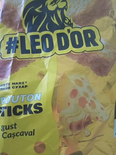 The image displays a close-up view of a crumpled yellow food packaging, likely for snacks. Dominating the upper portion of the package is a large graphic of a lion's head in black with yellow outlines. Below the lion, the hashtag "#LEODOR" is prominently displayed in large, black, stylized letters with a yellow outline. To the left of this text, some smaller text is visible, including what appears to be "SMETI MARI *" and "УПНИЙ СУХАР" in Cyrillic script. Further down, the words "ROUTON STICKS" are printed in large, silver-grey, sans-serif font. Below this, the words "gust" and "Casacaval" are written in a smaller, dark font, with "Casacaval" featuring a cedilla under the 'C'. The lower right section of the packaging shows an illustration of a slice of Swiss cheese with melting cheese. In the very bottom right corner, a line of very small text reads "UN EXEMPLU DE PREZENTARE A PRODUSULUI". The lighting is uneven, suggesting the package is indoors under artificial light, and the crumpled nature of the material indicates it's a flexible bag. There are no people or discernible environmental details beyond the product packaging itself. The location of Cotiujenii Mari, Moldova, is not depicted or suggested in the image content.