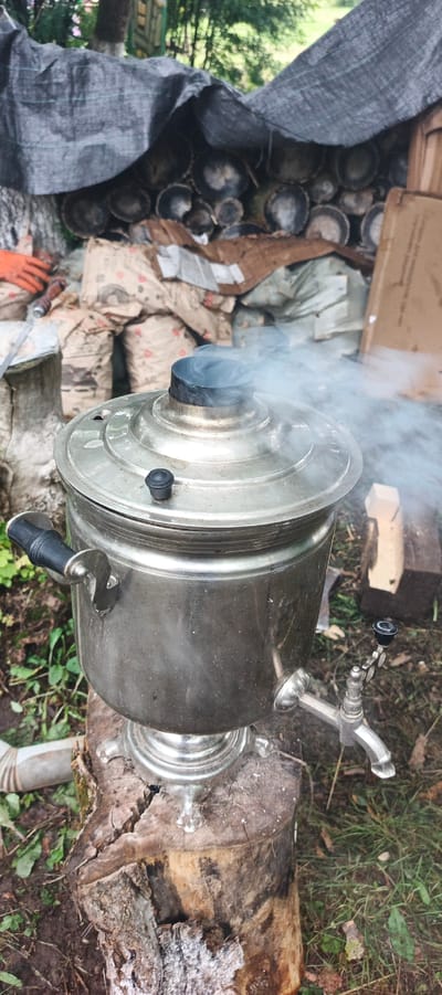 The image depicts an outdoor scene, possibly in Старые Быги, Russia. A traditional Russian samovar, made of silver-colored metal, is actively boiling and emitting steam from its top. The samovar is situated on a rough-hewn wooden stump. To the left of the samovar, an orange glove and part of a tool are visible, suggesting a person was recently working or is about to work. In the background, a pile of logs is covered by a dark tarp. There are also several large bags, some appearing to be sacks of materials, and a cardboard box. The ground is covered with dirt and green foliage, indicating a natural or semi-rural setting. The lighting suggests it is daytime.