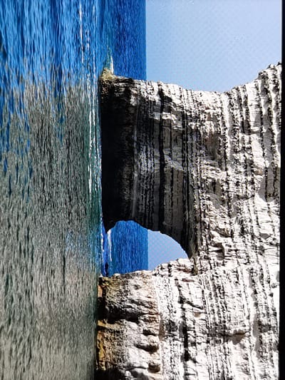 The image shows a dramatic view of the cliffs in Étretat, France, with the iconic Falaise d'Aval archway prominently featured. The deep blue of the ocean contrasts sharply with the white, layered rock formations of the cliffs. The water appears to be in motion, with visible ripples and waves, suggesting a dynamic seascape. The scale of the archway is emphasized by the immense cliffs surrounding it. The lighting suggests it might be daytime, with the sky a pale blue. The image is a close-up view, focusing on the texture of the rock and the water's surface.