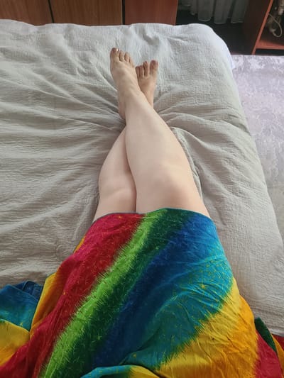 A person is lying on a bed, covered partially by a vibrantly colored, tie-dyed garment featuring stripes of red, orange, yellow, green, and blue. Their legs are crossed, and their feet are visible at the edge of the bed. The bed is covered with a light-colored, textured comforter. In the background, to the left, a wooden shelf unit is partially visible, with some items on its shelves. The scene appears to be indoors. No text is visible in the image.