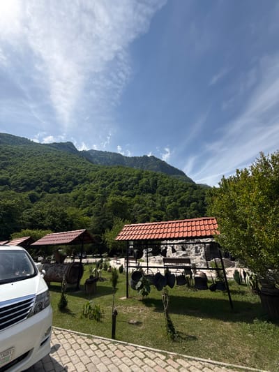This image captures a wide outdoor scene in Bzypta, Abkhazia, under a clear blue sky with sparse, wispy white clouds. Dominating the background are densely forested mountains, exhibiting various shades of green, suggesting a robust natural environment.

In the mid-ground, there are multiple open-sided structures with distinctive red-tiled roofs, likely pergolas or pavilions, indicating an outdoor establishment. One such structure is centrally located, and parts of others are visible on the left and right. Beneath one of these central structures, a large, horizontal wooden barrel, possibly decorative, is situated on a patch of green grass. Adjacent to it, to the right, a metal framework displays various dark-colored metal pots, pans, and buckets hanging decoratively. Behind these features, a light-colored, rocky or stone wall is visible. The ground is covered with well-maintained green grass and interspersed with small plants and shrubs.

In the immediate foreground, to the left, a portion of a white multi-purpose vehicle (MPV) or large sedan is parked on a light-colored, herringbone-patterned paved surface. The vehicle's front grille, headlights, and a partial view of its windshield are discernible. A license plate starting with "ABH" is partially visible on the lower left of the vehicle. To the far right, a lush green bush or small tree frames the edge of the scene.

The lighting is bright, indicative of a sunny day, with distinct shadows cast on the paved ground and under the structures. No individuals are visible in the image.