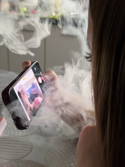 A close-up shot depicts a person's face and hand holding a smartphone engulfed in white smoke. Only the upper part of the face is visible, including the eyelashes and forehead beneath dark hair. The person's hand is holding a black smartphone, with the screen displaying a photograph. The scene is obscured by a thick cloud of white smoke, which partially obscures the background. The background appears to show some indistinct shapes and patterns, possibly fabric or furniture. The location, Port-Katon, Russia, is not discernible from the visual information provided.