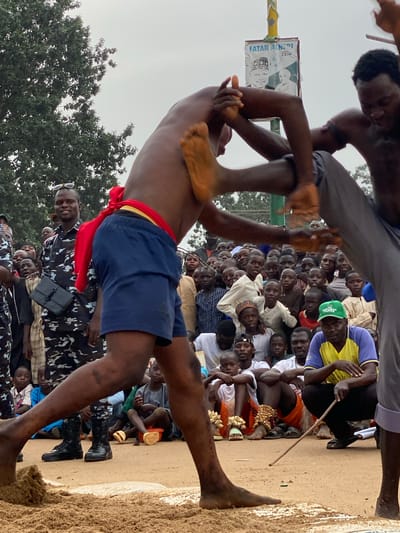 Two shirtless men are wrestling in a sand pit surrounded by a large crowd of onlookers in Kafanchan A, Nigeria. One wrestler wears blue shorts with a red sash, while the other wears grey pants. A poster hangs from a pole behind them.