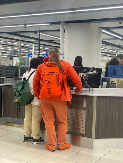 The image depicts an indoor scene within a modern public facility, likely an airport terminal, which could potentially be Gatwick Airport given its proximity to Crawley, United Kingdom.
In the foreground, two individuals are standing with their backs to the viewer at a grey-speckled counter with dark paneling below. The person on the right is wearing a monochrome orange outfit, including a long-sleeved jacket, trousers, and slip-on shoes. This individual has long, light brown hair tied in a ponytail with an orange elastic and is carrying a bright orange "Jordan" brand backpack featuring the Jumpman logo and "JOARDRA N" printed vertically in black text. Their right hand rests on the counter near a black computer monitor mounted on a silver articulated arm.
To the left of the person in orange, another individual wears a white long-sleeved shirt, beige trousers, and black sneakers with white soles. This person has dark, possibly braided, hair and is carrying a large, dark green backpack with a small brown patch on a lower pocket.
In the mid-ground, a large white cylindrical pillar partially obscures a third person. To the far right, a fourth person with dark hair is partially visible, leaning over a counter or machine.
The background shows a large, brightly lit space with a grid ceiling composed of dark beams and light grey panels, featuring numerous rectangular fluorescent light fixtures and several dome-shaped security cameras. To the far left, beyond the counter, a section resembling a security checkpoint or baggage screening area is visible with stacked grey trays, a green indicator light, and a blue sign. The floor is covered with light-colored square tiles.