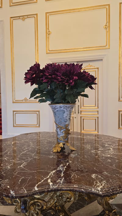 A floral arrangement of dark purple dahlias sits in a blue and white patterned vase on a polished brown marble table. The vase has gold accents and is supported by a golden, leaf-shaped stand. Behind the table, a wall features ornate gold trim outlining rectangular panels. A white door with gold hardware is visible in the background. The image appears to be taken indoors, possibly in a well-decorated room in Agios Epiktitos, Northern Cyprus.