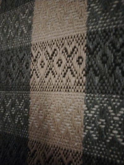 The image displays a close-up, slightly blurred view of a woven textile with a plaid pattern. The textile features alternating blocks of color and pattern. One set of blocks appears to be a neutral beige or cream color, with a textured weave. The other set of blocks is a darker grey or blue-grey, also with a textured weave, and incorporates a geometric diamond-shaped pattern in a lighter color. The pattern is intricate and appears to be a woven design rather than printed. The lighting suggests the textile is indoors, possibly a blanket or a rug. There are no people or identifiable environmental features visible in the image, and therefore, it cannot be definitively located in Stepnoe, Russia.