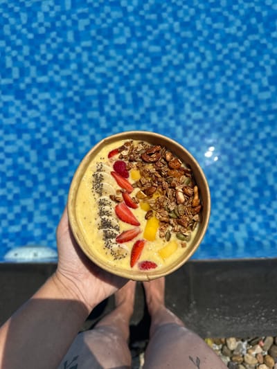 A bowl of smoothie decorated with sliced strawberries, banana, and mango, topped with granola and chia seeds, held by a hand near a swimming pool. The pool has small, blue mosaic tiles. Part of a person's legs are visible in the background next to the pool's edge. The smoothie bowl has a light yellow base and a brown paper-like container.