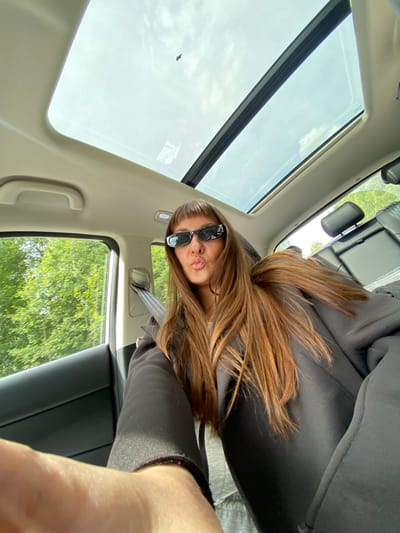 A woman with long auburn hair and bangs is sitting in the passenger seat of a car, holding up her phone to take a selfie. She is wearing black sunglasses and a black hoodie. The car has a sunroof open, revealing a cloudy sky. Trees and greenery are visible through the car window, suggesting a rural or suburban setting. The image is taken from a low angle, looking up at the woman and the car's interior. There is no identifiable location in the city of Гавриловка, Russia.