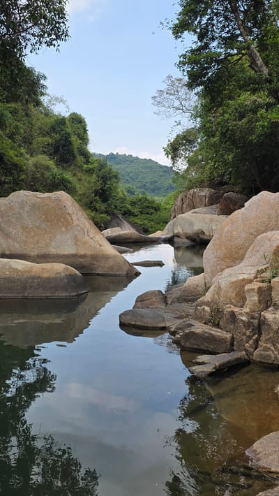 The image captures a tranquil scene in the city of Nam Ninh Hoa, Vietnam. Large, smooth rocks dominate the foreground, leading to a calm body of water, likely a stream or a small pond. The rocks reflect in the water, creating a mirror-like effect. Lush greenery covers the hillside in the background, with various trees adding to the natural beauty. The sky is visible in the upper left corner, suggesting a bright, daytime setting. No people are present in the view.