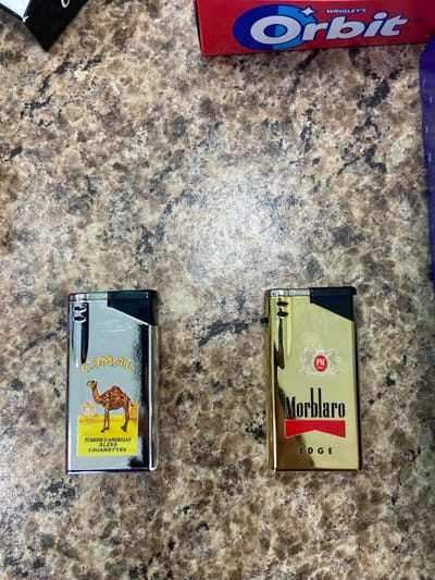 This is an overhead shot of two cigarette lighters and a box of Orbit gum placed on a speckled, marble-like countertop. The subjects are the lighters and gum, with the countertop serving as the setting.

The top lighter is gold with a black accent and features the branding "Morblaro EDGE" in red and black text. A circular logo with "PM INC." is also visible on the lighter.

Below the gold lighter is a silver lighter with a black accent. It displays the "Camel" brand logo, which includes a camel silhouette and text that reads "TURKISH & AMERICAN BLEND CIGARETTES."

To the left of the lighters is a red and blue box of "Orbit" gum, with the brand name prominently displayed in white and blue lettering.

There are no people visible, and no specific activity or event is depicted beyond the arrangement of the objects. The lighting suggests an indoor setting, likely during the daytime, with no indication of weather conditions or specific time of day. The location context of Yegorlykskaya, Russia, is provided, but there are no visual cues in the image to confirm this.