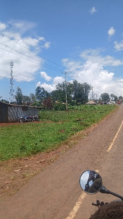 The image displays an outdoor scene captured from a motorcycle's perspective in Boochi Tendere ward, Kenya, under a bright blue sky with scattered white clouds. The foreground features the upper portion of a motorcycle's round, chrome-framed rearview mirror, reflecting a person wearing a brown, textured garment and part of their hand on the handlebars, along with a portion of the sky and road.

The main subject is a reddish-brown asphalt road, possibly a rural or semi-urban thoroughfare, marked with a faded yellow line on its right edge, curving slightly to the right and extending into the background. To the immediate left of the road is a strip of reddish soil, followed by an expanse of dense green ground vegetation and scattered taller plants.

Further to the left, a long, low building constructed with corrugated metal siding, likely galvanized iron, is visible. Parked directly in front of this building are approximately four motorcycles, some exhibiting blue and white accents. Beyond this structure and the vegetation, a cluster of trees with dark green foliage stands. In the far left background, a tall, lattice-structured communication tower (cell tower) equipped with multiple antennae rises prominently. Overhead, multiple power lines stretch across the frame, supported by utility poles visible further down the road.

Along the right side of the road, more green vegetation and trees are present. In the distant background, to the right of the road's curve, several vehicles, including what appear to be white vans or cars, and a group of people are visible, indicating some activity or gathering. A structure with a conical or thatched roof is partially discernible among the trees in the distance on the right.
