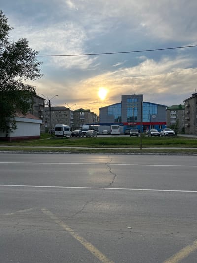 The image depicts a street scene in Uray, Russia. A multi-lane asphalt road with white lane markings stretches into the distance, showing some cracking and wear. A grassy strip separates the road from a row of parked white vans or small trucks. Beyond the vehicles are several buildings, including a modern blue and grey building with glass windows bearing the sign "МАГНИТ" in red lettering. Adjacent to this building are older, multi-story residential buildings. The sky is partly cloudy with the sun visible, creating a bright glare. A power line is visible running diagonally across the upper left portion of the image. The lighting suggests either early morning or late afternoon. A tree with green foliage is present in the lower left corner of the image.