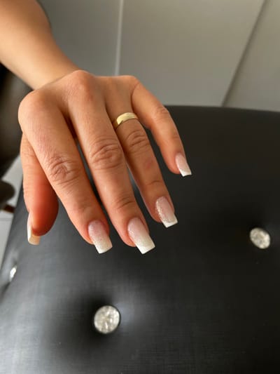 The image displays a close-up, eye-level perspective of a light-skinned human right hand, resting on a black, tufted, upholstered surface. A plain gold-colored band is present on the ring finger. All five visible fingernails are manicured in a square or squoval 'French manicure' style, featuring a glittery, translucent pink or nude base coat that transitions into opaque white tips. The dark upholstered surface exhibits two distinct, circular, reflective decorative studs. A lighter, off-white background element is visible in the upper-left quadrant behind the hand. The scene depicts an indoor setting, consistent with a professional nail salon or similar personal care establishment in Anápolis, Brazil.