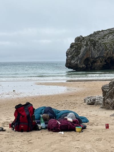 Two individuals are sleeping on a sandy beach near the town of Llanes, Spain. The scene includes various pieces of camping equipment, such as backpacks, sleeping bags, and a yellow bowl. A red can of soda is visible to the right of the sleeping figures. The ocean and a large rock formation are in the background. The sky is overcast.