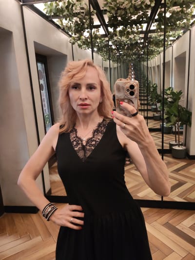 A woman with blonde hair, wearing a black dress with lace trim, takes a selfie in a room with mirrored walls and ceiling. She is wearing several bracelets on her left arm and rings on her fingers. The room has a wooden floor and potted plants visible in the reflections. The location is identified as Faliukovo, Russia.