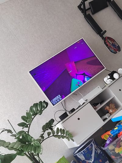 In a brightly lit, modern room with light gray textured flooring, a white entertainment unit sits against a wall. The unit has multiple shelves and drawers, some of which are open, revealing a collection of objects including toys, a decorative figurine, and a clear plastic bin filled with colorful building blocks. On top of the unit, a white gaming headset with black accents rests near a small white circular object. A television screen displays a vibrant purple and pink abstract image, likely from a video game, with on-screen text that includes "Bricks: 13574". A large green potted plant with glossy leaves is positioned to the left of the entertainment unit, its branches extending upwards. To the right of the television, a portion of a black and metal exercise machine is visible, positioned at an angle. The scene appears to be a living space or playroom.