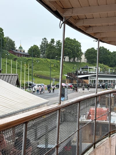 This detailed image depicts a rainy, overcast scene at the Lappeenranta harbor in Finland, looking towards the Linnoitus (Fortress) hill.
In the immediate foreground, a wooden railing with a grey metal chain-link fence extends diagonally from the lower left to the lower right, partially obscuring the view below. A wet, dark wooden deck is visible to the right of the railing. Behind the fence, several orange life vests are stacked. The upper right corner is occupied by a light brown, slatted underside of a roof structure.
The midground showcases a paved area or promenade. Approximately ten individuals are visible, mostly walking away from the viewer towards the left. Seven of these individuals are holding black or patterned umbrellas, indicating rain. One person is pushing a baby stroller. A black car is parked near the walking path, and a silver SUV is parked further right. To the right, a multi-level building complex with dark wood siding, white trim, and a flat roof terrace is prominent. A sign reading "Don Papa" is visible on the upper level. To the left of the paved area, a building with a light-colored, striped awning or roof is partially visible. Five tall, slender white poles are vertically aligned on a grassy slope, leading up the hill.
The background features a steep, verdant hill covered with numerous green trees. A traditional red-painted wooden building with a dark roof and white window frames is situated on the lower part of the hill. At the very top of the hill, the distinctive Orthodox Church of the Protection of the Mother of God (Pokrova Church) is visible, characterized by its light green dome and a golden cross atop a spire. The sky above is uniformly grey and overcast.