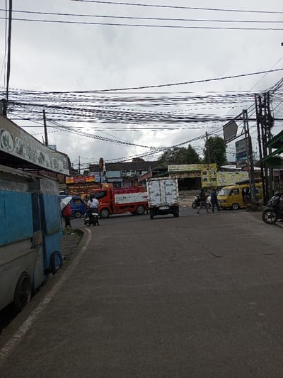 The scene depicts an active urban street in Melakudikadu, India, under an overcast sky. Numerous entangled black electrical and communication wires span horizontally across the upper half of the frame, originating from utility poles on both sides of the street. The foreground features an asphalt road surface with minor cracks, curving slightly to the left.
On the left side of the foreground, a light blue and white utility vehicle or stall is parked partially on an unpaved shoulder. Further down the street on the left, a person holding a white umbrella stands near a small blue utility van. Immediately to their right, a person on a black motorcycle, wearing a white shirt and dark helmet, is stopped in the road, facing slightly right.
In the mid-ground, a red flatbed truck with a white cab, loaded with multiple cylindrical objects (possibly gas tanks), is positioned in the road, appearing to be turning or halted. Behind it, a white box truck is visible from the rear. To the right of these vehicles, multiple individuals (at least three) are observed walking across the street. Further right, a yellow minivan or microbus is parked or stopped with its side door open, and at least two people are near it. A black motorcycle is partially visible on the far right in the foreground.
The background shows a dense array of commercial buildings and shops lining the street, displaying various signs with text. Trees and foliage are visible behind the structures in the mid-distance. Multiple utility poles with crossbars support the overhead wiring grid throughout the scene.