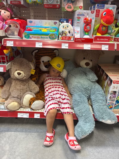 In Uuemõisa, Estonia, a young child is seated on a red shelf in a toy store, surrounded by large stuffed animals. The child wears a pink and white striped dress adorned with strawberries and pink Hello Kitty crocs. A yellow construction helmet is perched on their head. To the child's left are two giant teddy bears, one brown and one light blue, and to their right is a large stuffed tiger. The shelf displays various toys, including a "Pop-Up Animals" learning toy and plush dinosaurs from the "Baby Clementoni" brand. Price tags are visible on the shelf. The environment appears to be a well-lit retail space.