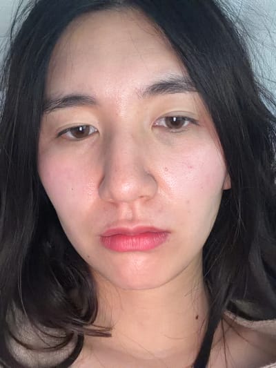 A close-up, portrait-style photograph shows a young woman with dark hair. Her skin tone is light, and she is wearing pink lipstick. Her eyes are brown. The background is a pale, neutral color. The photograph is taken from a slightly low angle. There is no visible indication of the location being Grimaud, France.