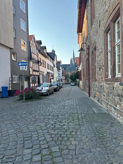The image depicts a narrow cobblestone street in Butzbach, Germany, lined with historic buildings. On the left, a modern grey building with several windows is visible, next to a traditional half-timbered building. A blue trash bin and a bush with red flowers are situated near the sidewalk. A street sign indicating a one-way street and a bicycle path is also present.

Several cars are parked along the right side of the street, which is bordered by a rustic stone building. In the distance, a church with spires is visible against a clear blue sky. The street is paved with dark gray cobblestones, with small patches of grass growing between them. The overall scene suggests a quiet, old European town.