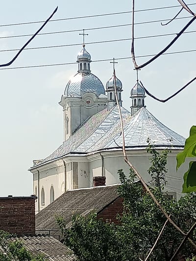 The image shows a white church with silver domes and crosses, partially obscured by tree branches and power lines. The church appears to be in the city of Yampil, Ukraine. The foreground consists of rooftops, vegetation, and the aforementioned branches and wires. The sky is clear and light blue.