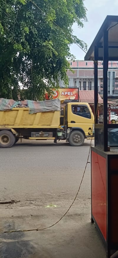 A yellow dump truck is driving on a paved road in what appears to be a town or village in Gampongteungoh, Indonesia. The truck is covered with a tarp, suggesting it is carrying a load. The setting is somewhat cluttered, with trees and buildings visible in the background. To the right of the frame, a red and black structure, possibly a food stall or kiosk, is partially visible. The lighting suggests it is daytime, and the sky appears overcast. A wire or rope is hanging down from the structure on the right, crossing over the road. A billboard advertising "APEL" with images of apples is visible behind the truck, indicating a possible local business or product. The overall scene depicts a typical street view in a populated area.