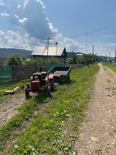 A bright, sunny day unfolds in Новомуллакаево, Russia, with fluffy white clouds dotting a vibrant blue sky. The scene is dominated by a rural, dirt road that winds through a village. In the foreground, to the left of the road, sits a small, red tractor with a hitch and an empty teal-colored trailer. The tractor appears weathered, with visible rust and a slightly worn appearance. Beside the tractor, a small pile of wooden planks rests on the gravelly ground.
A green wooden fence runs along the left side of the road, separating the properties. Beyond the fence, a yellow and blue house with a corrugated metal roof stands out, its details suggesting a traditional rural dwelling. More houses and trees are visible further down the road, stretching into the distance, with a gentle, tree-covered hill forming the backdrop. Power lines crisscross the sky above the houses, supported by utility poles. The overall impression is one of a peaceful, working village on a pleasant summer day.
