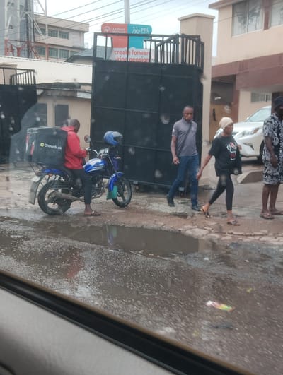 The image shows a street scene in Alagbede, Nigeria. In the foreground, a man on a blue motorcycle with a black "Chowdeck" delivery box is parked on a wet, muddy road. The rider is wearing a red jacket and dark pants. A blue helmet rests on the back of the motorcycle.

In the background, several people are walking. A man in a grey t-shirt and blue jeans is walking towards the right, with a bag slung over his shoulder. To his right, a woman wearing a black t-shirt, black leggings, and a headscarf is walking. Further to the right, part of a person in a patterned outfit is visible.

Behind the people, a large black metal gate stands, with a building visible through it. A sign on a pole near the gate reads "LEADWAY PENSIOn" and another section of text is partially visible with the words "create forwar kind pension". A white car is parked near the building.

The road appears to be wet, with puddles reflecting the scene, suggesting recent rain. The lighting indicates it is daytime, possibly overcast. The overall atmosphere seems to be that of a busy street in an urban or semi-urban area.
