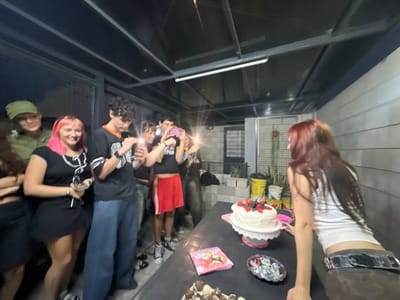 The image captures an outdoor gathering at night, under a covered patio structure, situated in a residential area of Quilmes Oeste, Argentina. Approximately eight young individuals are present.
In the immediate foreground, a dark rectangular table displays a white two-layer cake adorned with strawberries and blueberries, resting on a white pedestal stand. Adjacent to the cake are a pink decorative item, a small black dish containing colorful objects, and a pink wrapper featuring cartoon characters. A portion of another dark-colored dessert is visible in the bottom-left corner of the table.
On the right side of the table, a female with long, reddish-brown hair is seen from behind, leaning forward. She wears a white tank top and dark pants secured with a broad belt.
To the left of the table, a group of seven individuals is gathered, facing towards the cake. From left to right: a partially visible person; an individual wearing an olive-green hat and jacket; a female with bright pink hair, smiling and holding a smartphone, dressed in a black top with white detailing and a black skirt; a male with dark, curly hair, wearing a black and orange jersey and loose blue pants, holding a smartphone; a female with dark hair, wearing a dark top and red shorts, holding a smartphone with an active flash directed forward; and two other individuals partially visible behind her, one also holding a smartphone with an active flash.
The environment includes a dark overhead structure with exposed metal beams and artificial lighting fixtures. The background consists of a light-colored concrete block wall on the right and, further back, a building or enclosure with a window/door featuring horizontal bars. Potted plants, a yellow bucket, and various items are visible along the base of these structures.