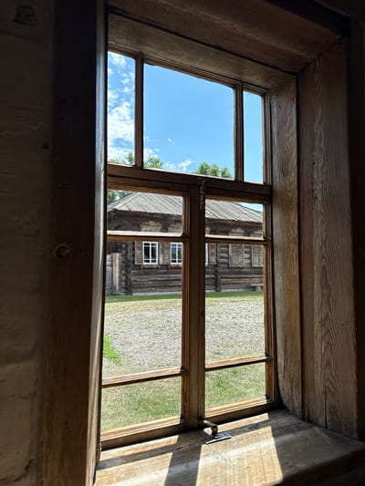 The image shows a view through a wooden-framed window onto an outdoor scene. The window frame itself is made of rough-hewn wood and is divided into several panes by horizontal and vertical wooden mullions. Sunlight streams in through the window, casting distinct shadows on the wooden windowsill and the floor beyond.
Through the window, a rustic wooden building with a pitched roof is visible. The building appears to be constructed from logs, a common architectural style in rural Russia. It has several windows with white panes and wooden shutters. The area directly in front of the building is covered with light-colored gravel and patches of green grass. In the background, trees are visible against a bright blue sky with scattered white clouds.
This scene is likely from the Shushenskoye museum-reserve, a historic site in Shushenskoye, Russia, dedicated to preserving the memory of Vladimir Lenin's exile there. The architectural style of the building and the rustic setting are consistent with the historical buildings found in such reserves. The image captures a moment of tranquility, with the warm sunlight illuminating the scene.